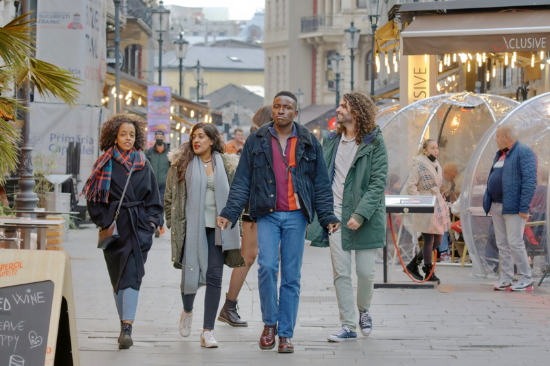 1 - A diverse group of four friends walking together down a lively, cobblestone pedestrian street lined with outdoor dining areas and clear-dome igloo seating